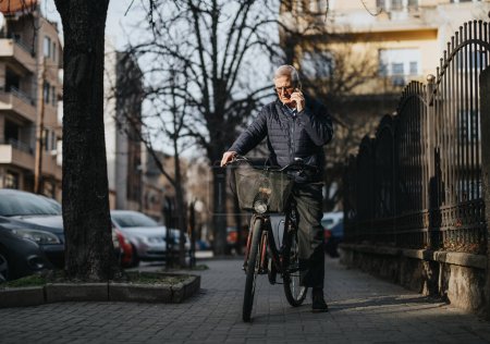 Senior man on phone while walking with bicycle on urban street.