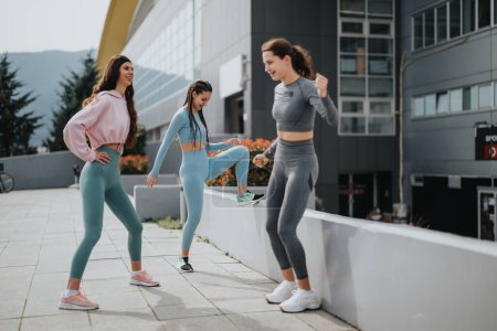 Drei junge Frauen dehnen sich vor einem Workout, in sportlichen Outfits, vor urbaner Kulisse.