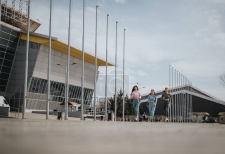 Tres mujeres jóvenes corriendo juntas cerca de un edificio moderno, promoviendo la aptitud y la amistad.