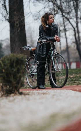 Stilvolle und selbstbewusste Frau, möglicherweise Geschäftsfrau, mit Kopfhörer um den Hals, an einem Oldtimer-Fahrrad in einer ruhigen Parklandschaft stehend.