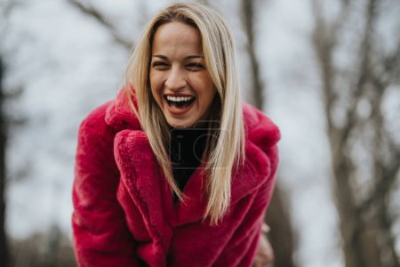 A joyful young blonde woman wearing a striking pink coat is laughing heartily in an outdoor, winter setting.