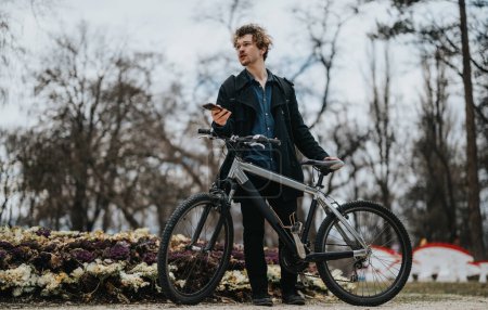 Un joven socio de negocios se detiene durante un paseo en bicicleta para trabajar de forma remota usando su teléfono inteligente en un entorno de parque.