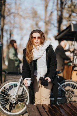Junge Frau mit Sonnenbrille genießt einen sonnigen Wintertag im Freien und hält eine Kaffeetasse in der Nähe einer Parkbank.