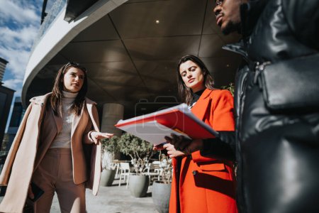 Two young businesswomen in stylish winter coats engaged in a serious discussion with paperwork outside a contemporary office on a sunny day.