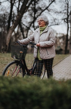 Reife Rentnerin steht mit ihrem Fahrrad in einer ruhigen Parklandschaft und blickt in die Ferne.