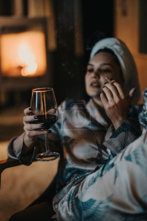 A serene moment as a woman with a head towel unwinds with wine and a cigarette by a warm fireplace.