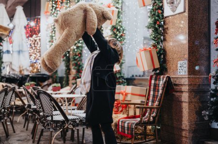 Eine festliche Outdoor-Szene, in der jemand neben einem schön dekorierten Café einen großen Teddybär und Geschenkboxen in der Hand hält.