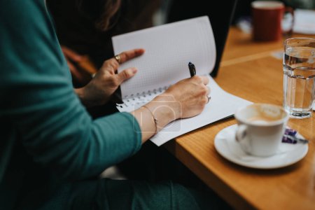 Nahaufnahme eines Geschäftsmannes, der neben einer Tasse Kaffee und einem Glas Wasser in ein Notizbuch schreibt