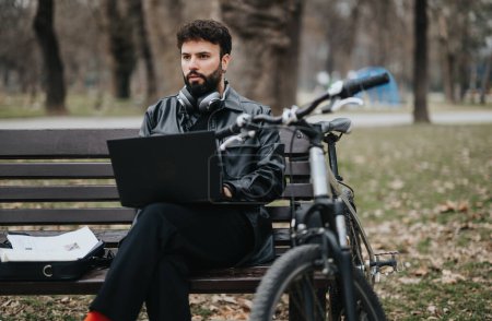 Focused businessman in casual attire working remotely on a laptop while sitting on a park bench, alongside his bicycle in an urban setting.