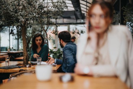 Deux entrepreneurs concentrés discutant de stratégies commerciales autour d'un café dans un cadre confortable café avec une femme floue ayant un appel d'affaires assis à l'avant.