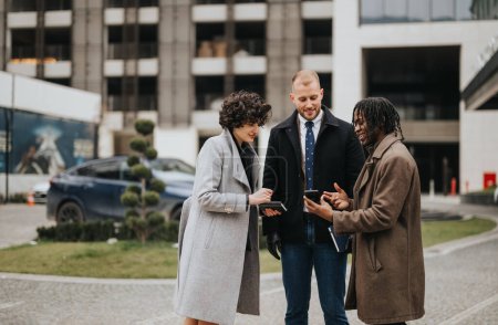 Drei junge multikulturelle Geschäftskollegen diskutieren vor einem Bürogebäude und tauschen sich über digitale Geräte aus.