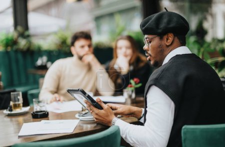 Konzentrierter afrikanischer Geschäftsmann mit Baskenmütze und digitalem Tablet bei einem lockeren Geschäftstreffen an einem Stand mit Kollegen im Outdoor-Café.