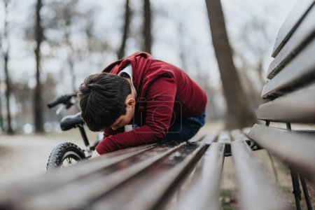 A tired boy takes a rest on a park bench with his bicycle parked nearby, capturing a moment of exhaustion