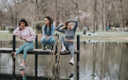 Ein aufrichtiger Moment, in dem drei junge Frauen auf einem Steg mit Blick auf das ruhige Wasser in einer ruhigen Parklandschaft entspannen.