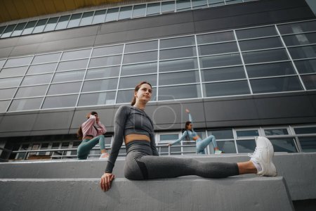 Active young women ready for exercise, stretching in an urban setting with modern architecture surroundings.