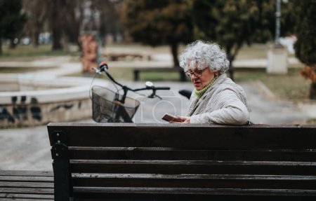 Un jubilado maduro y femenino disfruta del tiempo libre en un parque, sentado con una bicicleta y mirando su teléfono inteligente.
