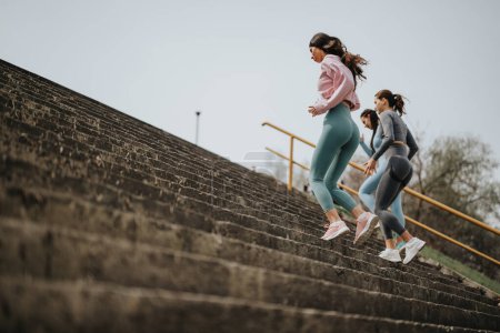 Three women in workout clothes running up steps outdoors, focused on fitness and healthy lifestyle.
