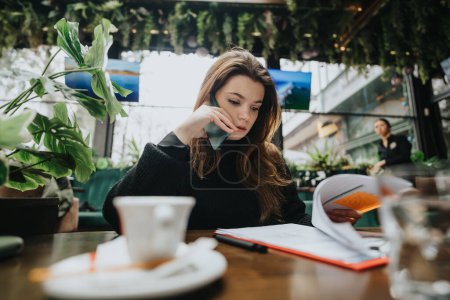 A young female entrepreneur is deeply focused while making a phone call and reviewing notes at a bustling urban coffee shop.
