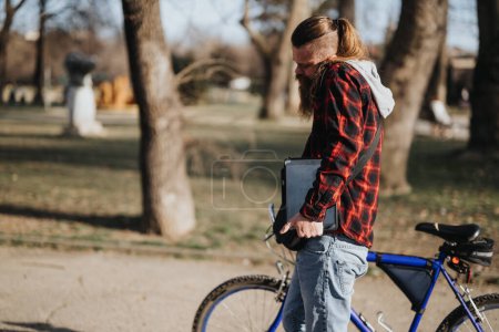 Un homme d'affaires barbu en tenue décontractée tenant un ordinateur portable debout à côté de son vélo dans un parc urbain, illustrant la flexibilité du travail à distance.