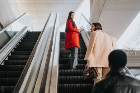 Two stylish women share a moment of connection on an escalator. One in a vibrant red coat, smiling over her shoulder at her friend below.