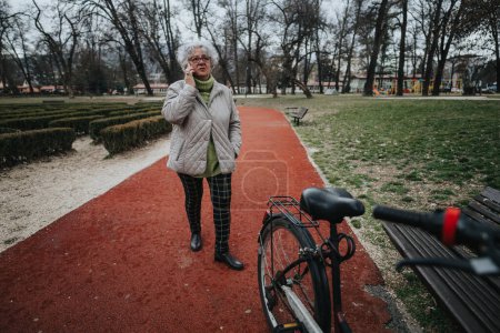 Mature female retiree standing next to her bicycle in a park, embodying active and healthy lifestyle choices.