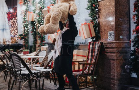 An individual with a large teddy bear and a gift box walking past a beautifully decorated outdoor cafe with Christmas lights and trees.