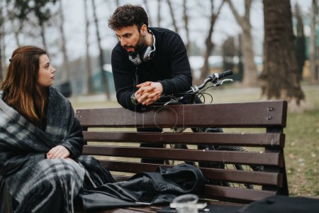 A candid moment depicting a man with headphones and a young woman wrapped in a blanket engaging in a deep conversation on a park bench, with a bicycle and coffee beside them.