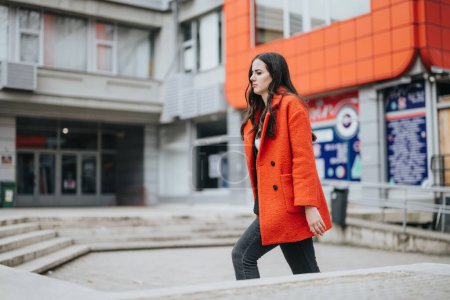 A motivated businesswoman walks confidently in an urban setting, wearing a stylish red coat and black pants.