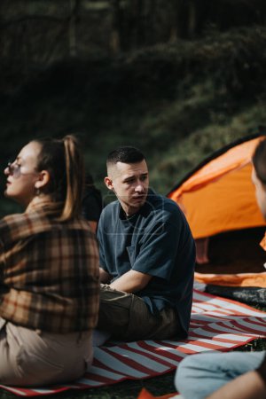 Group of friends seated on a picnic blanket during a camping adventure surrounded by greenery and a tent.