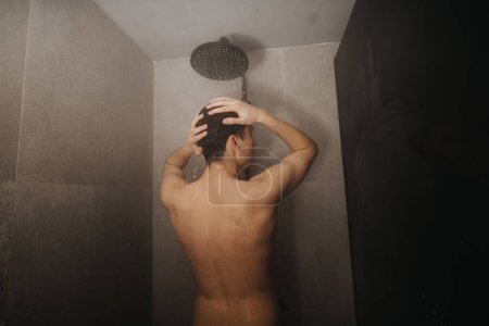 Rückansicht eines jungen Mannes, der sich in einem modernen Badezimmer unter einem Duschkopf die Haare spült