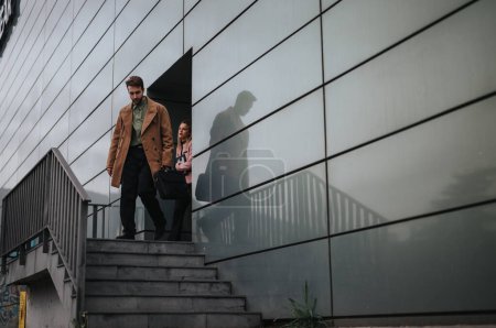 Dos jóvenes profesionales en atuendo casual de negocios bajando los escalones fuera de una estructura de oficinas contemporánea.