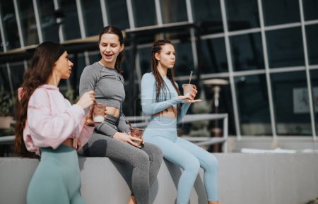 Drei Frauen in sportlicher Kleidung sitzen draußen, schlürfen Smoothies und genießen nach einer Fitnesseinheit Gesellschaft..
