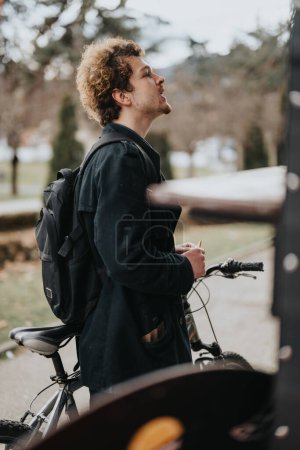 A professional businessman with a backpack standing next to a bicycle, taking a break outside in an urban park setting.