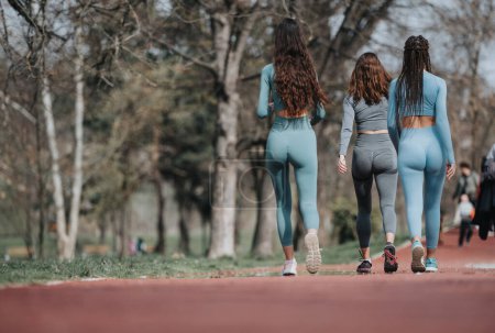 Eine Gruppe von drei Freundinnen in sportlicher Kleidung spaziert an einem sonnigen Tag im Park zusammen und symbolisiert Gesundheit und Kameradschaft.