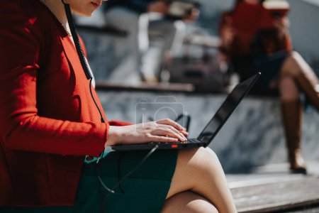 Femme professionnelle en blazer rouge utilisant un ordinateur portable pour les affaires en plein air avec des collègues en arrière-plan.