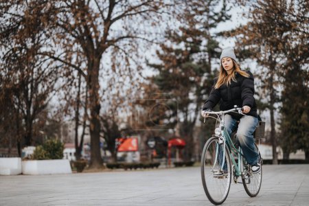 Casually dressed young woman with a beanie cycling through a serene park with autumn leaves, exuding a relaxed and peaceful vibe.