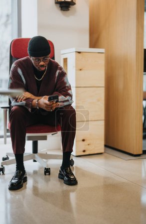 Ein stilvoller junger Erwachsener ist in sein Smartphone vertieft und sitzt gemütlich in einem Bürostuhl, der zeitgenössische Arbeitskleidung präsentiert..