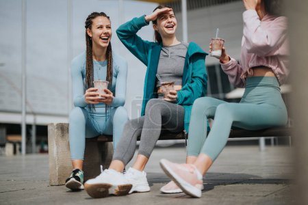Trois jeunes femmes assises à l'extérieur, prenant une pause avec des boissons rafraîchissantes après une séance de fitness.