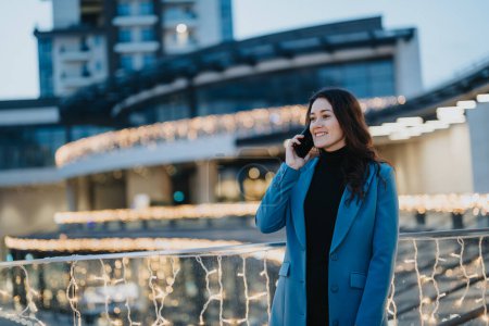 Smiling businesswoman in a blue coat using a mobile phone on a city street adorned with sparkling Christmas lights.