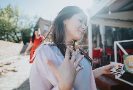 Eine zufriedene Frau entspannt sich und genießt ihren Kaffee an einem hellen Tag vor einem Café, während sich im Hintergrund eine andere Person nähert.