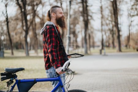 Stilvoller bärtiger Geschäftsmann mit Fahrrad, der in einem Stadtpark Pause macht, während er an einem sonnigen Tag aus der Ferne arbeitet.