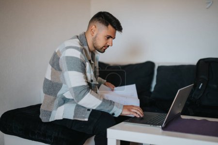 Konzentrierter junger Geschäftsmann begutachtet Papierkram, während er mit seinem Laptop auf einem Hocker in einem gut beleuchteten Raum sitzt