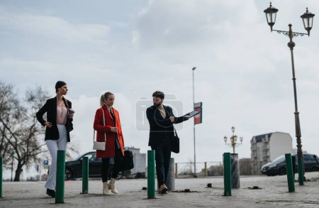 Eine Gruppe von drei Freunden hängt an einem trüben Tag in der Stadt herum und plaudert, während sie auf öffentliche Verkehrsmittel warten.