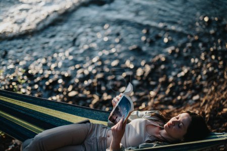 Niña despreocupada relajándose con un libro en una hamaca al aire libre, disfrutando de la naturaleza y la puesta de sol junto a un lago tranquilo.