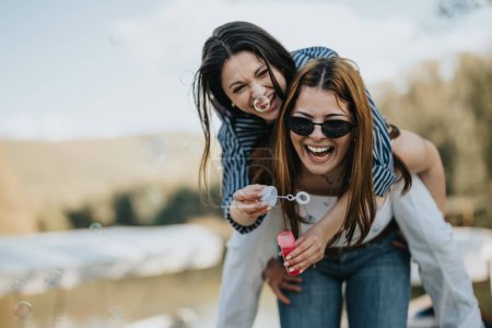 Dos jóvenes felices disfrutando de un momento lúdico con burbujas de jabón al aire libre cerca de un lago, expresando alegría y amistad.