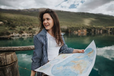 A happy young woman sightseeing at a beautiful lake, holding a map, dressed in a striped jacket, surrounded by mountains and clear waters.