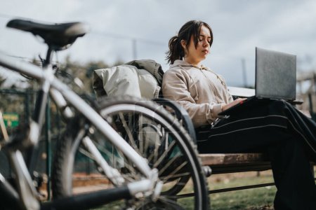 Ein junges Mädchen sitzt entspannt auf einer Parkbank, in ihren Laptop vertieft, ihr Fahrrad neben sich, und verkörpert Freizeit und digitalen Nomaden-Lebensstil..