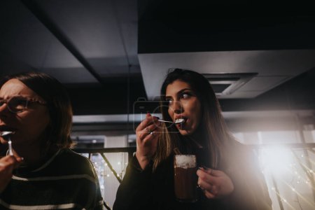 Dos mujeres comparten un momento con bebidas en un bar con poca luz y luces brillantes en el fondo, que representan la amistad y la relajación.