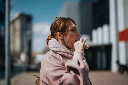 Eine junge Berufsfrau in lässiger pinkfarbener Jacke isst einen Imbiss, während sie an einem hellen Tag in einer städtischen Umgebung steht.