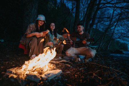 Three young adults relax by a campfire in the woods, playing guitar and enjoying drinks, surrounded by trees and the dark blue twilight.